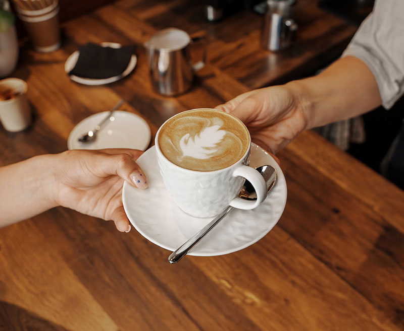 A barista hands over a beautifully crafted cappuccino with latte art to a customer at a wooden cafe counter